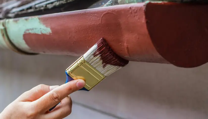 Ręka maluje pędzlem rynny, odnawiając stare malowanie aluminium.