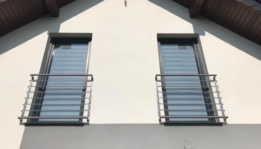 Nowoczesny montaż balkonu francuskiego na białej elewacji domu. Dwa okna z balustradami ze stali nierdzewnej.