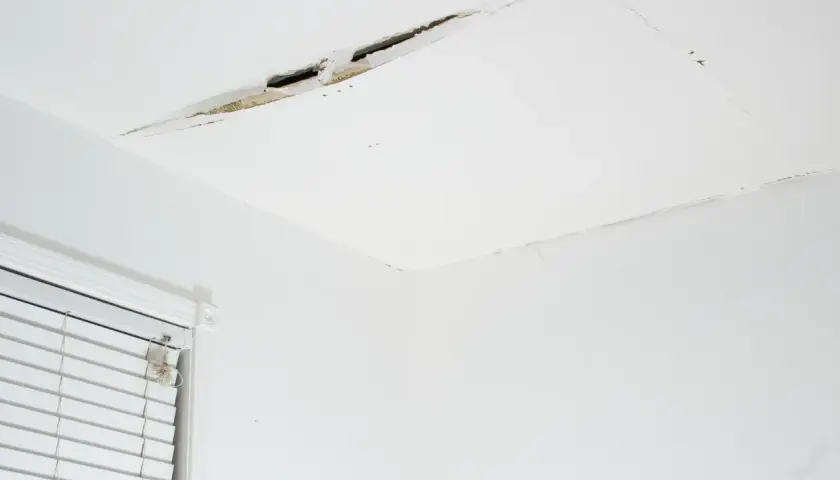 Widoczne pęknięcia na białym suficie, świadczące o problemach z konstrukcją lub wilgocią. Obok okno z białymi żaluzjami.