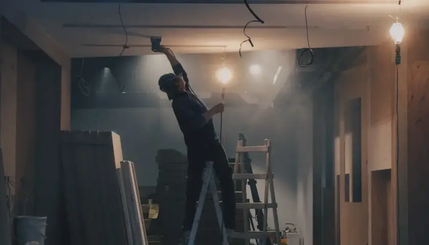 Budowa wnętrza, pracownik na drabinie montuje sufit, w tle materiały budowlane i lampy.