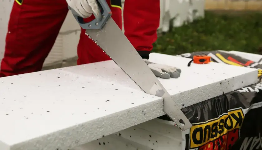 Ręce w rękawiczkach tną styropian zwykłą piłą ręczną. To prosty sposób, czym ciąć styropian.