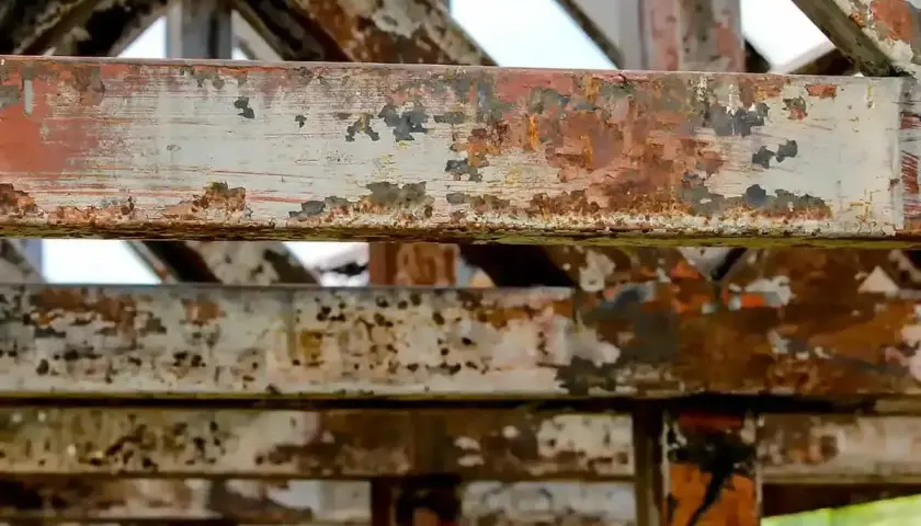 Metalowe belki konstrukcji pokryte łuszczącą się farbą i rdzą. Widać ślady postępującej korozji, która niszczy materiał.