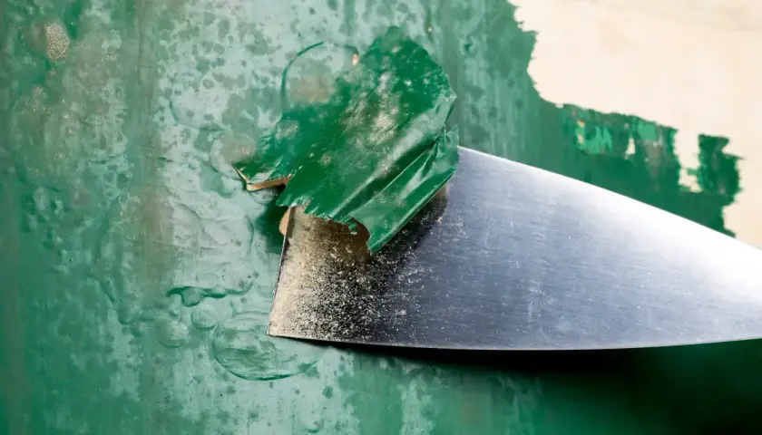 Metalowa szpachelka zdziera starą, zieloną farbę, odsłaniając podkład.