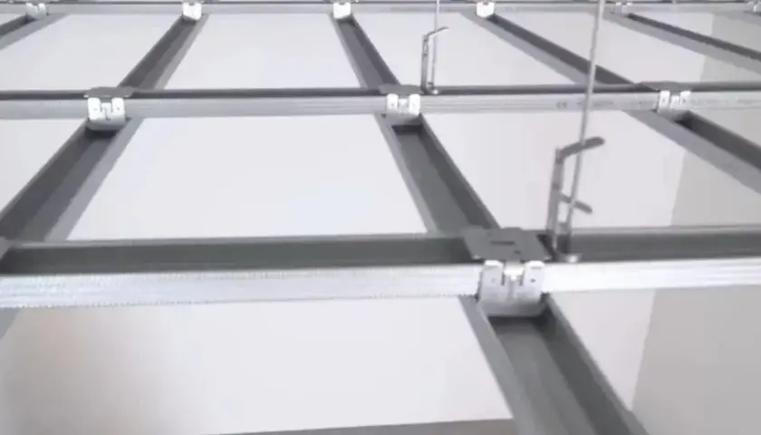 Sufit podwieszany krzyżowy z białymi panelami i metalową konstrukcją. Widać system profili, który tworzy siatkę, a nie pojedyncze belki.