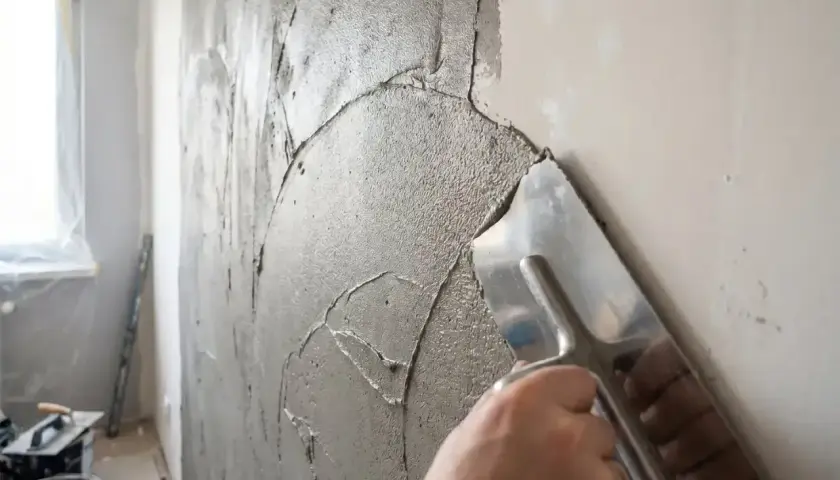 Ręka z kielnią nakłada grubą warstwę masy naprawczej na ścianę, tworząc nierówną fakturę.