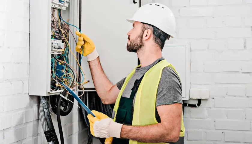 Elektryk w kasku i kamizelce odblaskowej pracuje przy skrzynce z przewodami. Zastanawia się, ile za punkt elektryczny zapłaci klient.