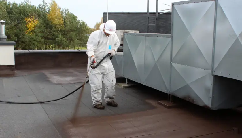 Pracownik w kombinezonie ochronnym wykonuje renowację papy termozgrzewalnej na dachu, natryskując preparat.