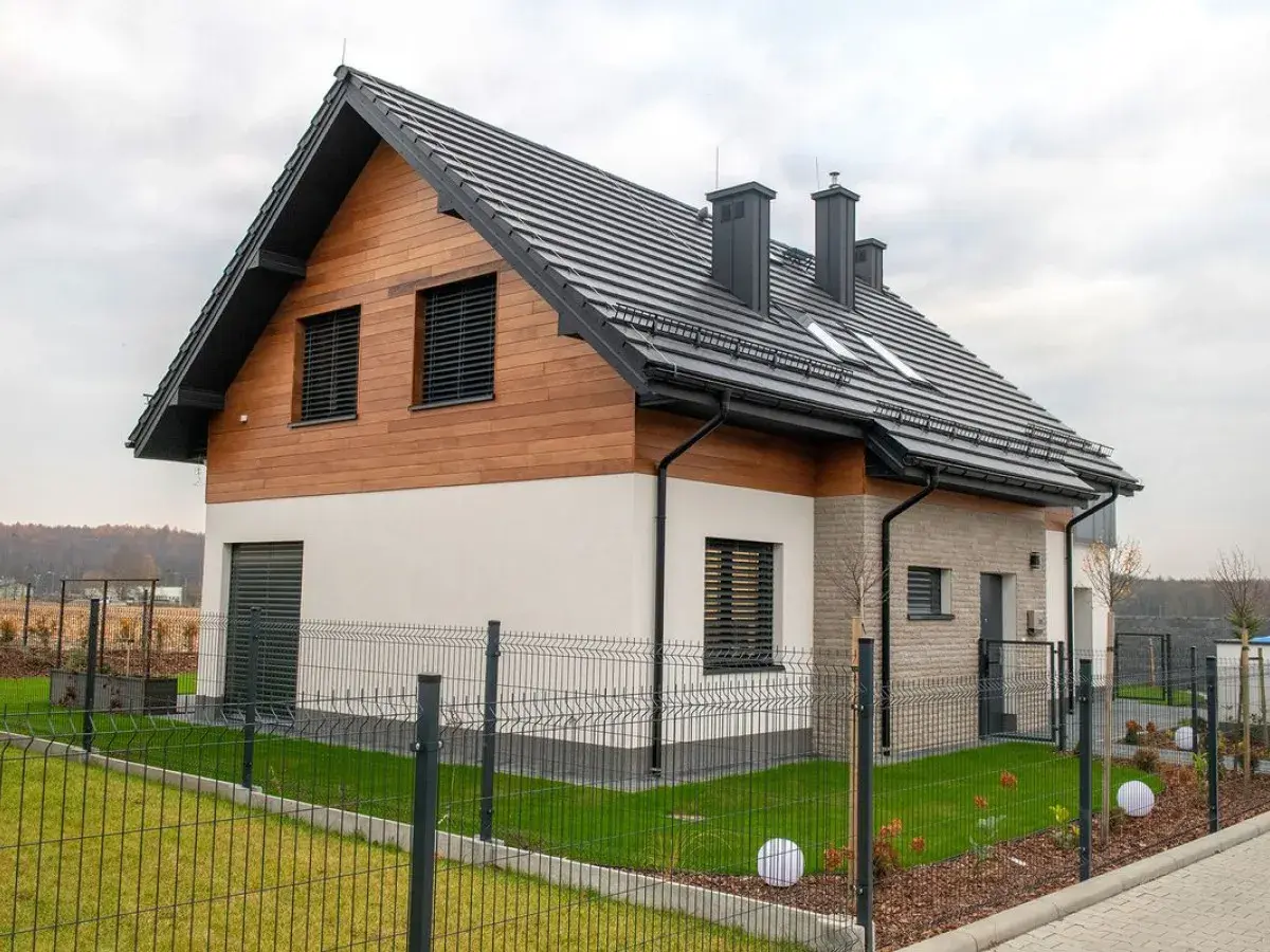 Nowoczesny dom z drewnianą elewacją i czarnym, dwuspadowym dachem. Ogrodzony teren z zadbanym trawnikiem i ozdobnymi kulami.
