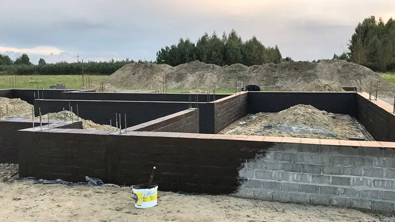 Fundament domu w budowie, ściany fundamentowe pokryte ciemną masą izolacyjną. Trwa izolacja powłokowa.