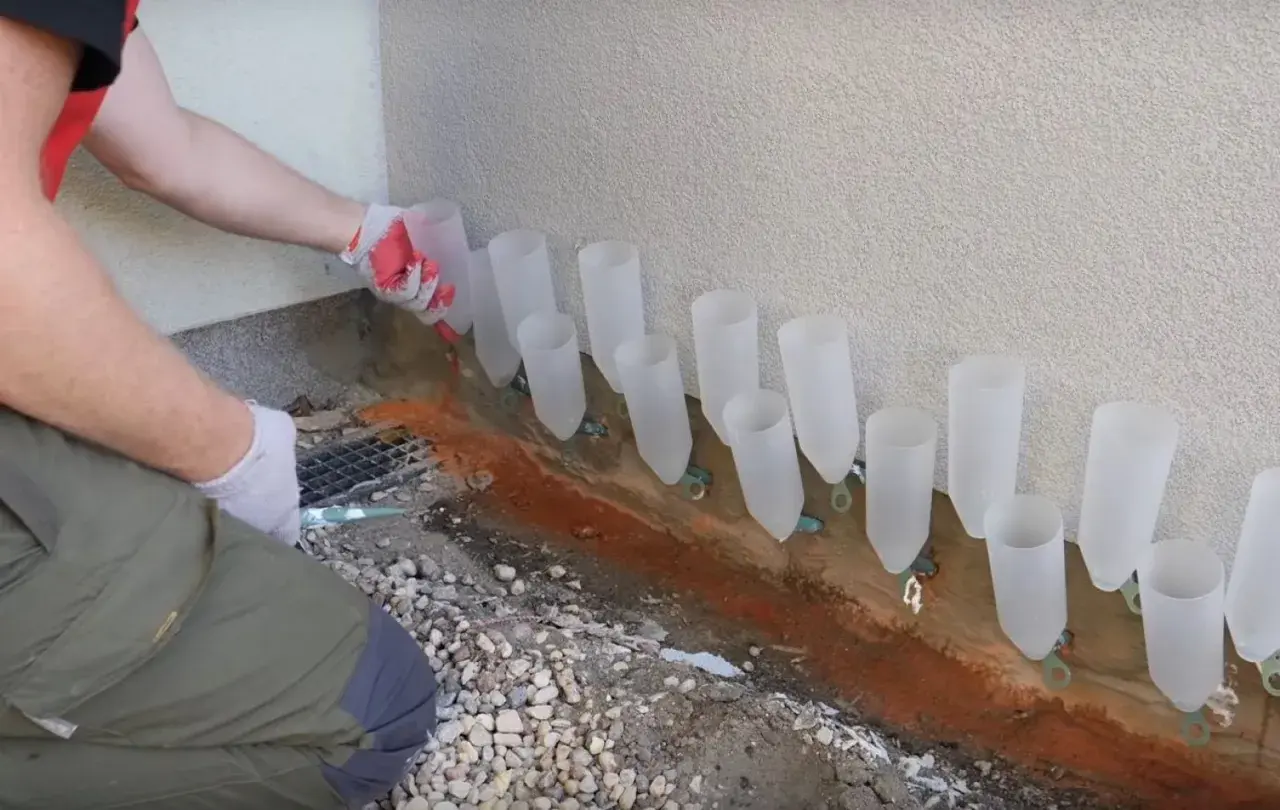 Pracownik w rękawiczkach montuje system do osuszania murów, umieszczając plastikowe rurki wzdłuż ściany.