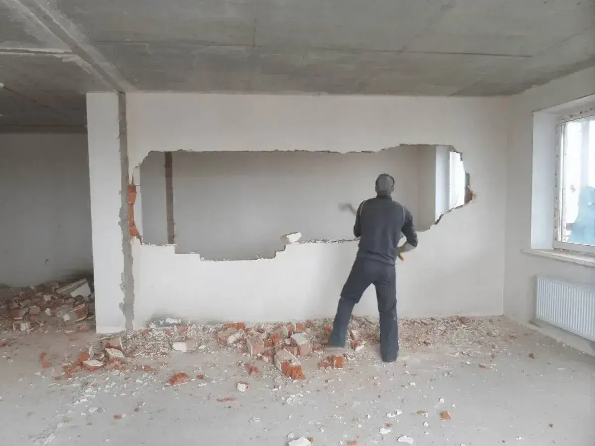 Mężczyzna młotem wyburza białe ściany, tworząc duży otwór. Wokół leżą gruzy.