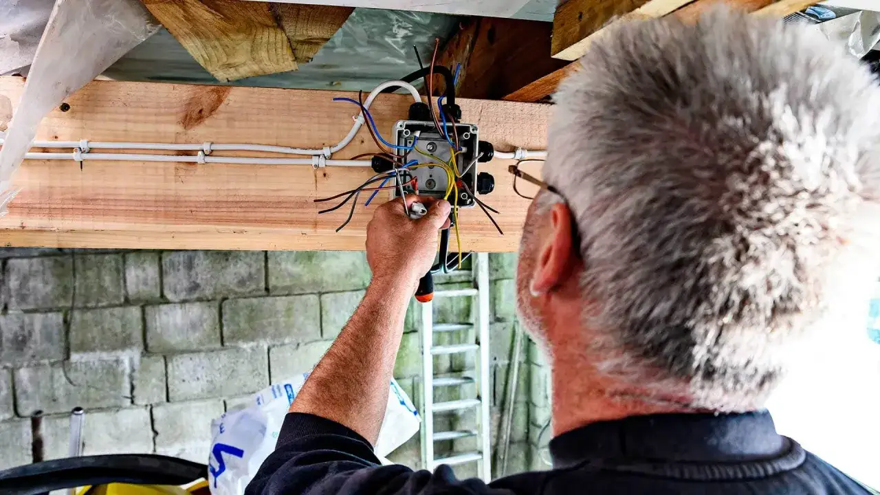 Elektryk montuje instalację elektryczną natynkową na drewnianej belce. Widać skrzynkę z przewodami i narzędzia.