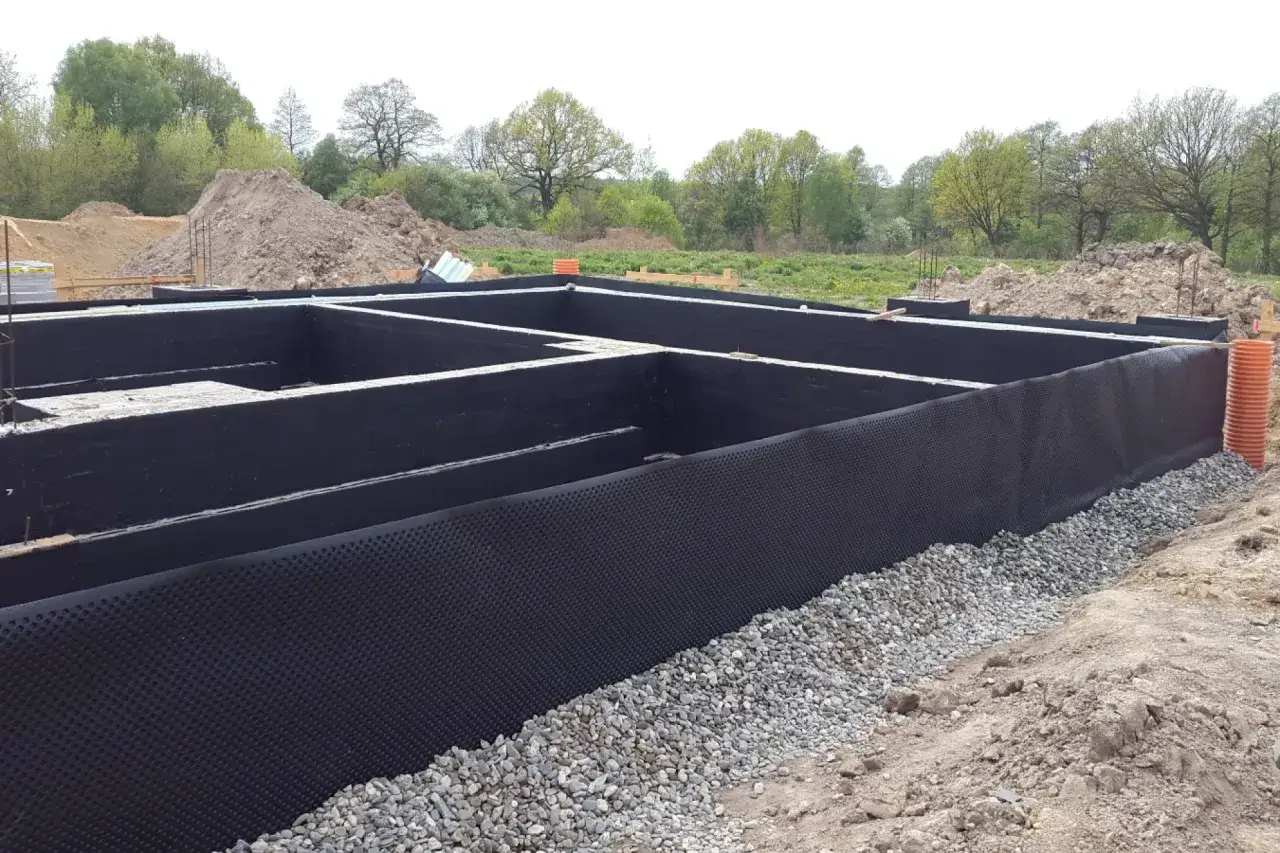 Fundament domu z czarną izolacją powłokową, otoczony żwirem i otwartym terenem budowy.