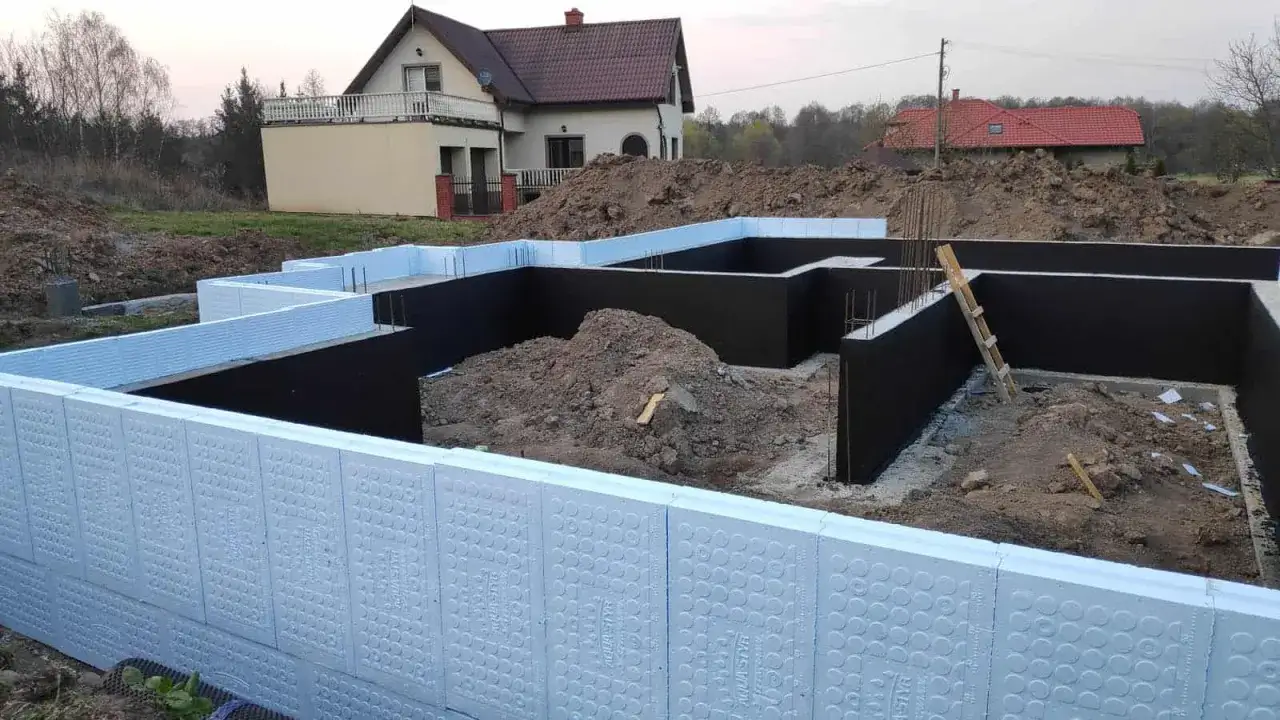 Fundament domu w budowie, z widocznymi ścianami z czarnej hydroizolacji i białym polistyrenem.