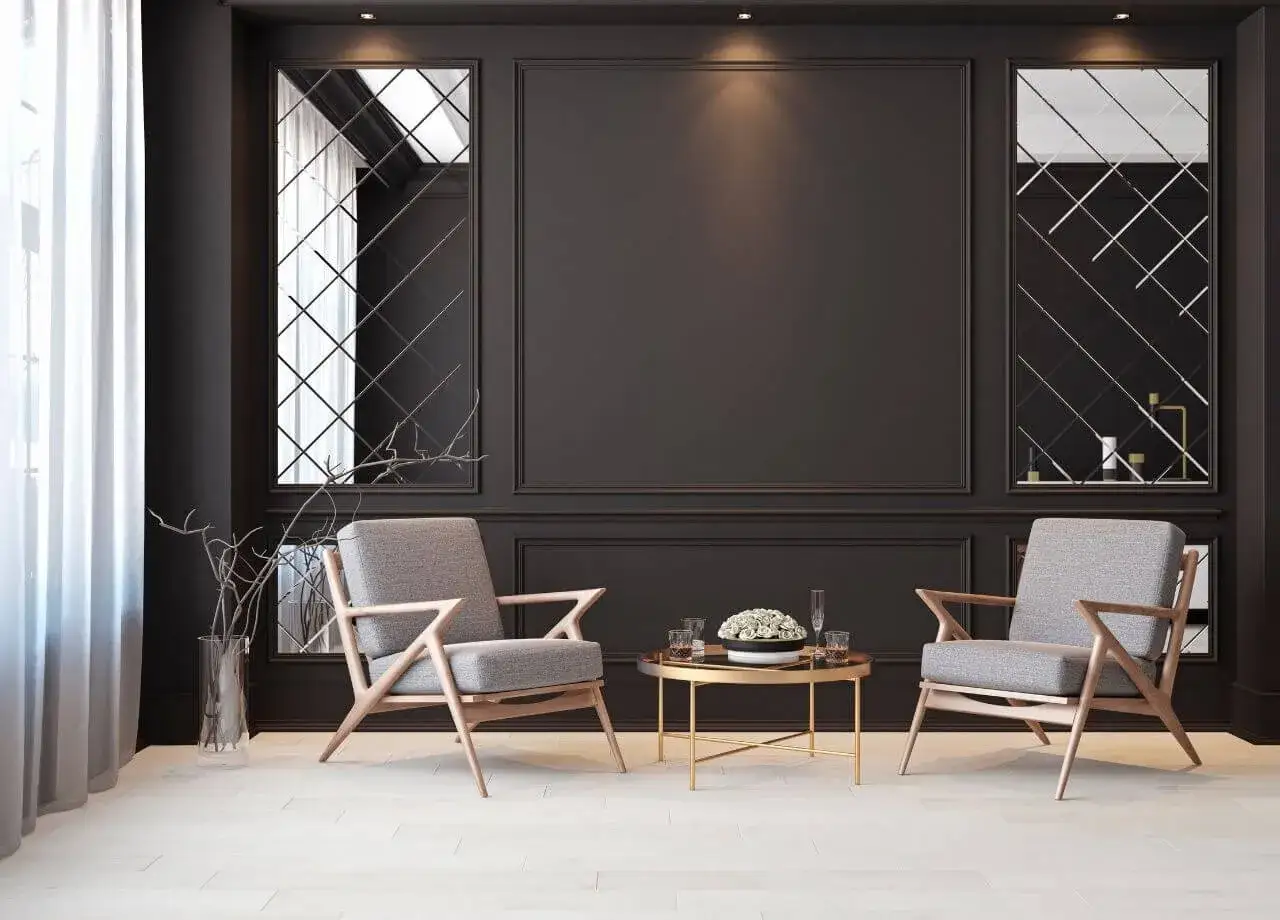 Elegancki salon z dwoma szarymi fotelami i złotym stolikiem. Ciemna ściana z lustrami tworzy intrygującą atmosferę, idealną do rozważenia, którą ścianę w pokoju pomalować na ciemniejszy kolor.