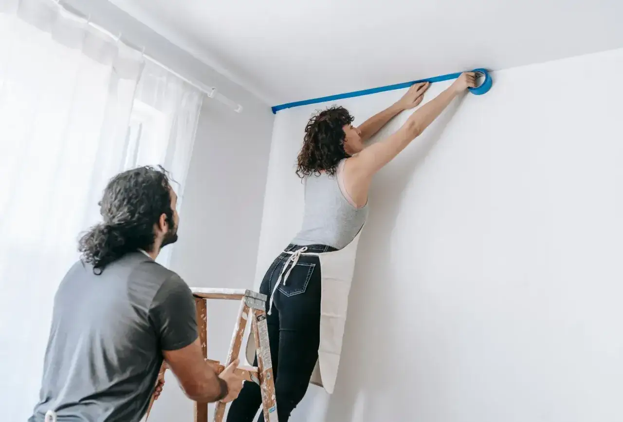 Para przygotowuje się do malowania sufitu, oklejając krawędzie taśmą.