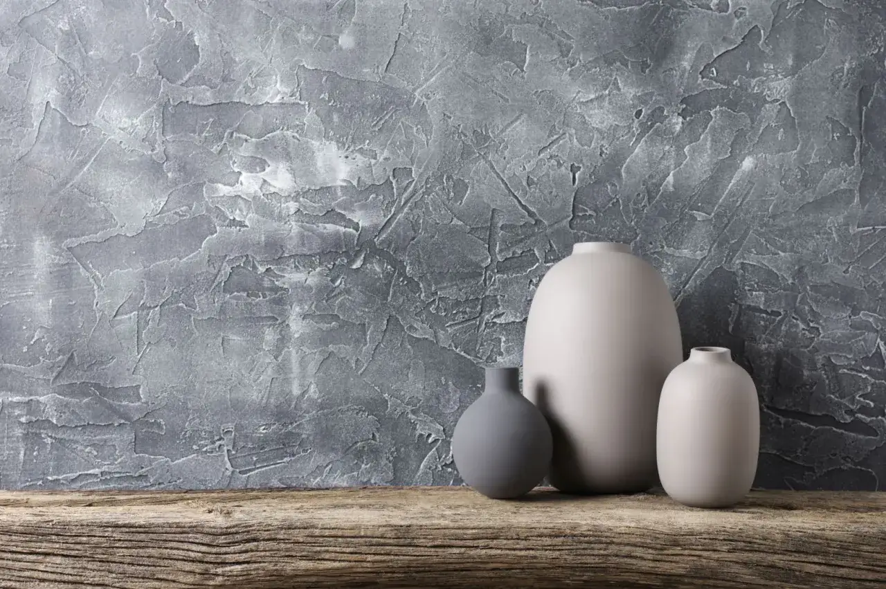 Trzy ceramiczne wazony na drewnianej półce, na tle ściany z surowymi tynkami strukturalnymi, tworzącymi ciekawe wzory.