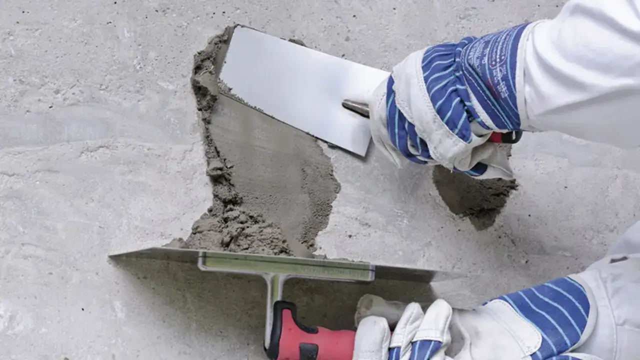 Dłonie w rękawicach nakładają masę naprawczą na betonową ścianę za pomocą kielni.